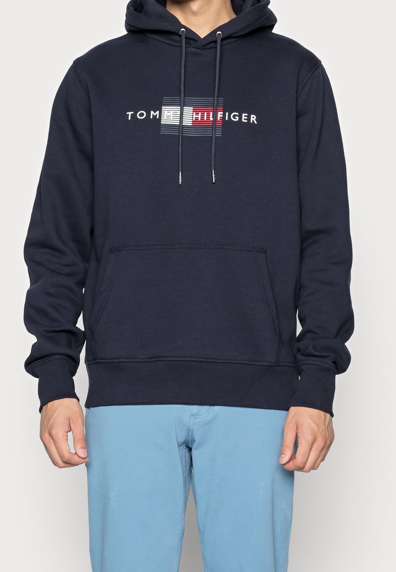 Personne portant un sweat à capuche bleu marine avec poche kangourou devant et le logo "Tommy Hilfiger" au-dessus d’un motif de drapeau rouge, blanc et bleu.