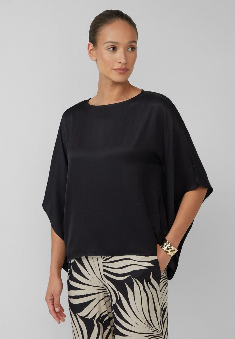 Haut en satin noir avec une coupe décontractée et des manches chauve-souris courtes. Encolure arrondie, texture lisse, associé à un pantalon à motifs beige et noir.