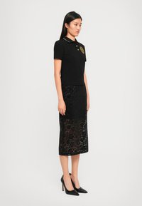 Black polo shirt with gold accents, paired with a black lace midi skirt featuring floral patterns. Model wears black pointed heels.