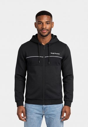 Uomo con felpa nera con cappuccio e zip Sergio Tacchini e jeans azzurri, in piedi davanti a uno sfondo bianco uniforme.