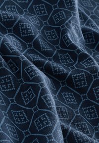 Temno modro blago z geometričnim vzorcem diamantov v svetlejši modri barvi, na katerem so vidne gube in tekstura pletenega materiala.