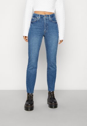 Osoba nosząca niebieskie dżinsy z wysokim stanem i surowymi wykończeniami oraz czarne platformowe buty na sznurowadła, w połączeniu z białym, fakturowanym długim topem typu crop.