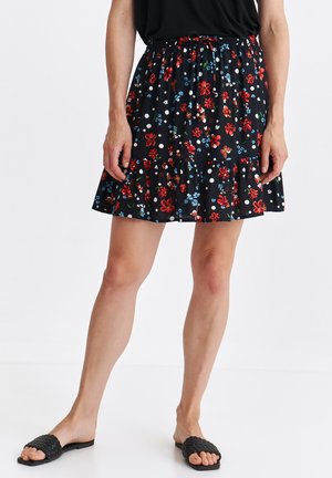 Czarna spódnica w kwiaty z czerwonymi i niebieskimi kwiatami oraz białymi kropkami. Elastyczny pas, warstwowy fason z falbaną na dole. Noszona z płaskimi czarnymi sandałami.