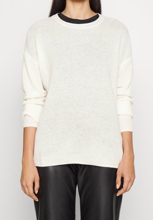 Femme portant un pull en maille blanche sur une chemise noire, associé à un pantalon en cuir noir, vue du cou aux hanches.