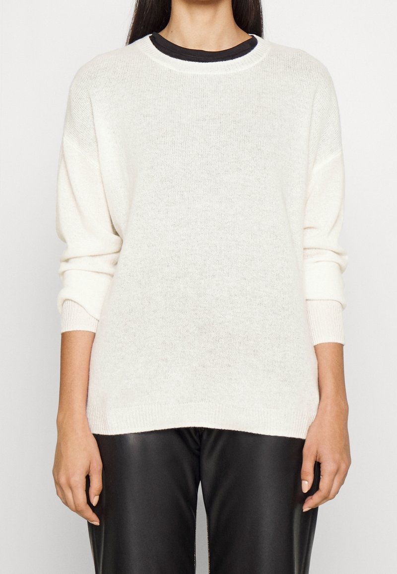 Femme portant un pull en maille blanche sur une chemise noire, associé à un pantalon en cuir noir, vue du cou aux hanches.