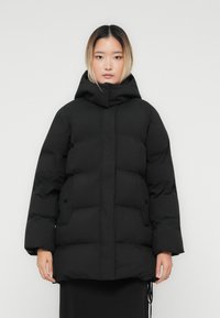 Junge Frau mit glattem Ombre-Haar, die eine schwarze gepolsterte Winterjacke mit Kapuze und Fronttaschen vor einem schlichten Hintergrund trägt.
