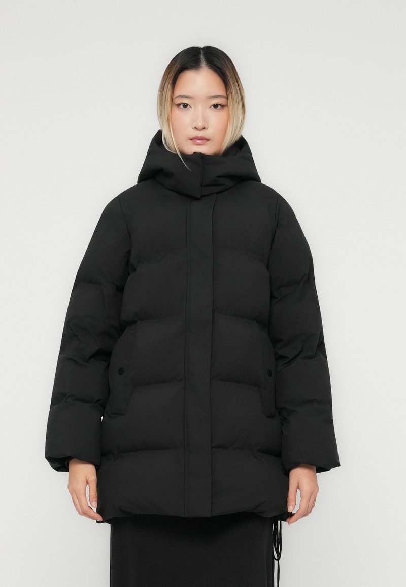 Junge Frau mit glattem Ombre-Haar, die eine schwarze gepolsterte Winterjacke mit Kapuze und Fronttaschen vor einem schlichten Hintergrund trägt.