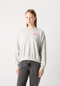Lichtgrijze sweatshirt met een ontspannen pasvorm, rib gebreide manchetten en tailleband. Bevat een roze logo. Draagt met donkergrijze jeans. Zachte textuur.