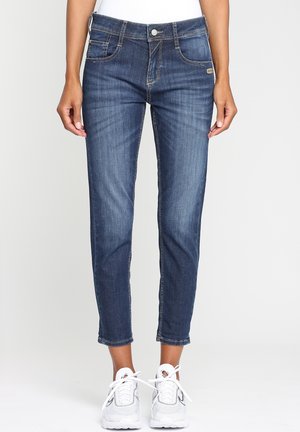 Femme portant un jean slim bleu foncé raccourci au-dessus des chevilles et des baskets blanches, debout devant un fond clair uni.