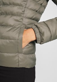 Oliwkowa, pikowana kurtka ocieplana z elastycznymi mankietami i kieszeniami po bokach. Materiał wydaje się błyszczący i gładki.