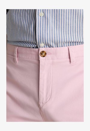 Šviesiai rožinės medvilninės kelnės su lygiu paviršiumi, turinčios rudos spalvos sagos užsegimą ir šonines kišenes, vilkimos su mėlynos ir baltos spalvų dryžuota palaidine.