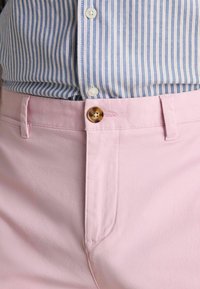 Lichtroze katoenen broek met een gladde textuur, voorzien van een bruine knoopsluiting en zijzakken, gedragen met een blauw-wit gestreept overhemd.