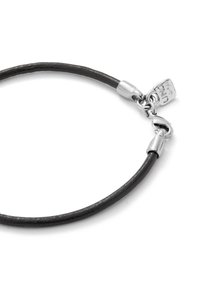 Bracelet en cuir noir avec une texture lisse, présentant des accessoires en métal argenté et un fermoir. Étiquette remarquablement gravée avec une marque.