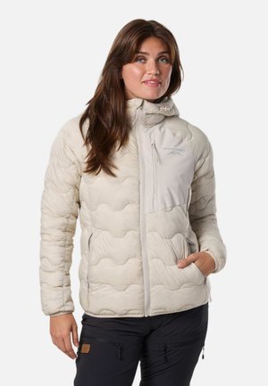 Une femme pose avec une veste à capuche matelassée couleur crème et un pantalon noir, sur un fond clair et uni.