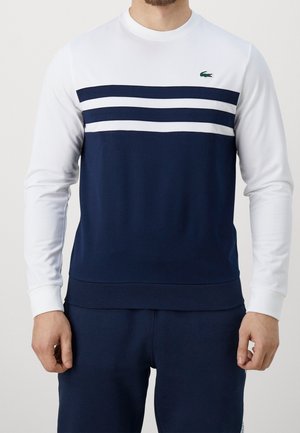 Mężczyzna ubrany w białą i granatową bluzę z długim rękawem z dwoma poziomymi granatowymi pasami na piersi oraz małe zielone logo.