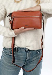 Borsa a tracolla in pelle marrone con chiusura a zip, accessori in metallo argentato e tracolla regolabile, appoggiata su un maglione lavorato a maglia color crema.