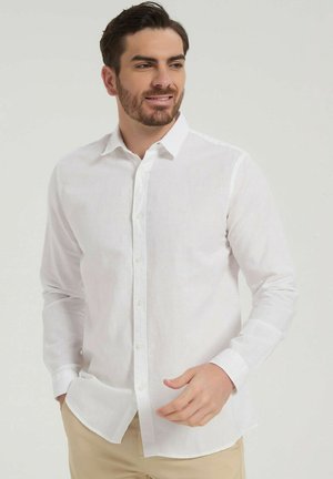 Homme avec une barbe taillée portant une chemise blanche à manches longues boutonnée et un pantalon beige, regardant sur le côté avec un léger sourire.
