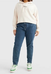 Hoodie cropped color rosa chiaro con cordoncini e testo "Jeans". Abbinato a jeans tapered in denim blu e sneakers verde-bianco.