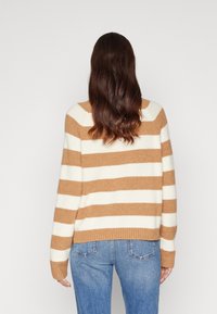 Beżowo-kremowy sweter w paski o luźnym kroju, ze ściągaczami na mankietach i dole, wykonany z miękkiego materiału, zestawiony z niebieskimi dżinsami.