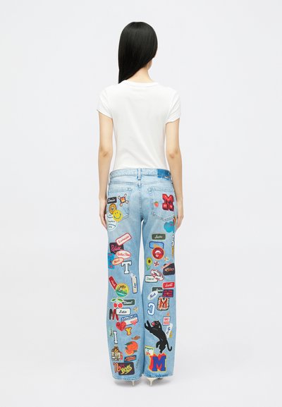 Jeans din denim de culoare albastră deschisă, prezentând diverse patch-uri colorate, inclusiv text și grafică, pe spatele fiecărei picioare. Asortate cu o cămașă albă simplă.
