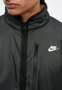 Černá bunda Nike s vysokým límcem, která má centrální zip a kapsu na zip. Materiál má hladkou texturu a lesklý povrch.
