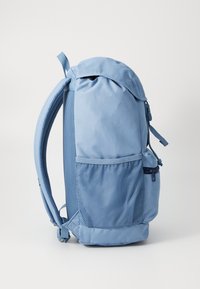 Light blue backpack with a rounded top, mesh side pocket, adjustable straps, and a zippered compartment. Smooth fabric with a textured accent.