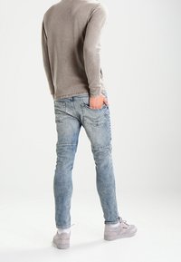 Chemise à manches longues gris clair, jean bleu déchiré avec une coupe ajustée, et baskets grises. Les jeans présentent un design à cinq poches et une texture décolorée.