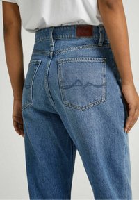 Blue denim jeans featuring a high waist, straight leg design, and back pockets with a subtle stitching pattern. Leather brand patch on waistband.