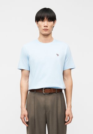 Άνδρας όρθιος, φοράει ανοιχτό γαλάζιο μπλουζάκι με μικρό λογότυπο ζέβρας, καφέ ζώνη και γκρι πιετέ παντελόνια σε απλό φόντο.