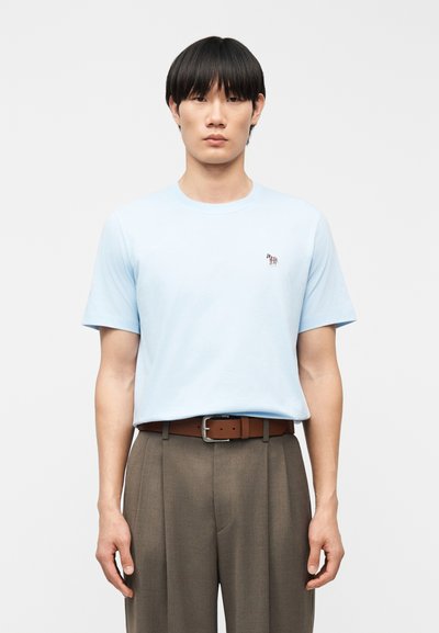 Hombre de pie, erguido, vistiendo una camiseta azul claro con un pequeño logo de cebra, cinturón marrón y pantalones grises plisados, sobre un fondo liso.