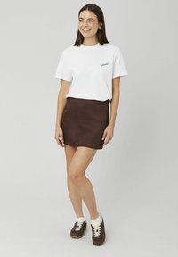 T-shirt en coton blanc avec le mot « please ! » en texte bordeaux, assorti à une mini-jupe en daim marron et des baskets foncées avec des accents blancs.