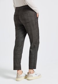 Persoon in donkergrijze geruite broek en witte sneakers, staande met handen in de zakken tegen een effen lichte achtergrond.