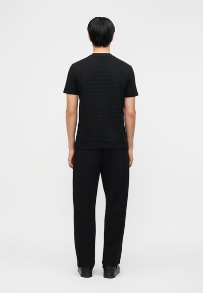 Άτομο όρθιο με την πλάτη προς τα πίσω, φοράει μαύρο πουκάμισο με κοντά μανίκια και υφή, μαύρο παντελόνι και μαύρα παπούτσια, μπροστά σε απλό φόντο.