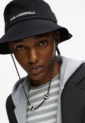 Czarna czapka typu bucket z logo "KARL LAGERFELD", noszona na szarym hoodie i w pasiastej koszuli, z koralikowym paskiem na szyi.