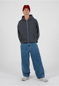 Grauer Hoodie mit Reißverschluss und Kapuze, kombiniert mit lockeren blauen Jeans und grauen Sneakers. Ein roter Strickmützen-Akzent vervollständigt den Look.