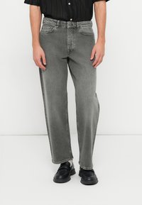 Pilkos džinsinės kelnės turi laisvą siluetą su šiek tiek siaurėjančiomis kojoms, penkių kišenių dizainą ir subtilų nudilimą. Pora dėvimi su juodais batais.