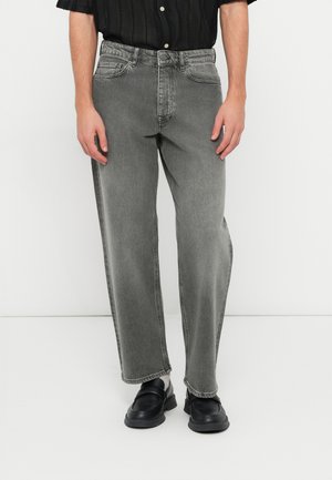 A szürke farmernadrág laza szabású, enyhén szűkülő lábbal, öt zsebes dizájnnal és diszkrét kopással rendelkezik. Fekete cipővel viselve.