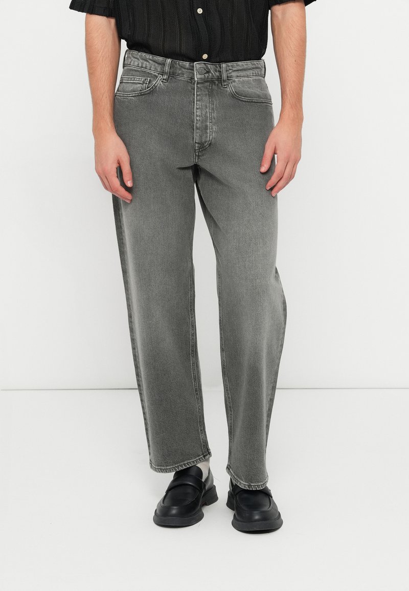 Pilkos džinsinės kelnės turi laisvą siluetą su šiek tiek siaurėjančiomis kojoms, penkių kišenių dizainą ir subtilų nudilimą. Pora dėvimi su juodais batais.