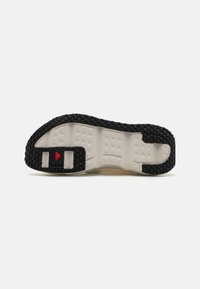 Semelle extérieure en caoutchouc noir avec une texture antidérapante, ornée d'un accent rouge central et de structures de soutien intégrées. Semelle intermédiaire crème visible au-dessus.