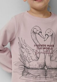 Laps, kes kannab roosa särki, millel on detailne luige illustratsioon ja tekst "Sõbrad muudavad maailma kaunimaks" ette.