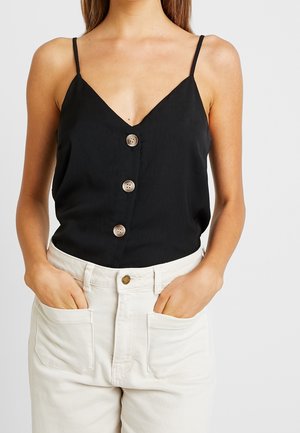 Mujer con una camiseta sin mangas negra con botones y pantalones de cintura alta color blanco roto con las manos en los bolsillos contra un fondo liso.
