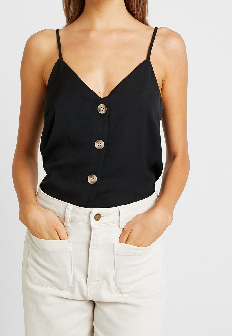 Femme portant un haut sans manches noir avec des boutons et un pantalon taille haute écru, les mains dans les poches, sur un fond uni.