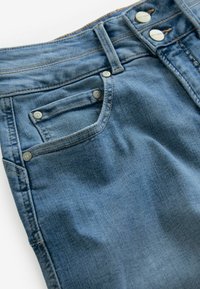 Denim džinsi vidēji zilos toņos ar gludu tekstūru, ar divām metāla pogām, mazu priekšējo kabatu un kontrastējošiem šuvju rakstiem.