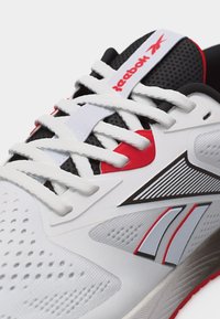 Λευκό και μαύρο αθλητικό παπούτσι Reebok με κόκκινες λεπτομέρειες, λευκά κορδόνια και επάνω μέρος από διαπνέον πλέγμα, κοντινή λήψη της μπροστινής πλευράς και του λογότυπου.