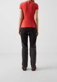 Rød t-shirt med korte ærmer, parret med sorte jeans, der har et lysere panel på benene. Model iført sorte sneakers med røde detaljer.