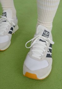 Adidas kedas ar baltu acs audumu, pelēku zamšu akcentiem, melnām svītrām un oranžu zoli. Kombinēti ar krēmas krāsas, ribotām zeķēm.