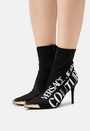 Bottes de cheville en tissu noir avec un bout pointu et une fermeture zippée. Présente des lettres blanches et un accent de talon métallique doré.