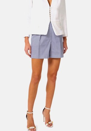 Donna che indossa una giacca bianca traforata, pantaloncini a righe verticali blu e bianche e sandali bianchi con tacco alto e cinturini alla caviglia.