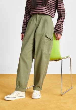 Osoba stojąca obok zielonego krzesła, ubrana w khaki bojówki, białe sneakersy i brązowo-czarną pasiastą koszulkę z długim rękawem.
