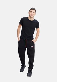 Camiseta negra y pantalones de chándal negros con un cordón de colores. Los pantalones de chándal cuentan con un logo blanco y una construcción sólida. El modelo está de pie de manera casual.